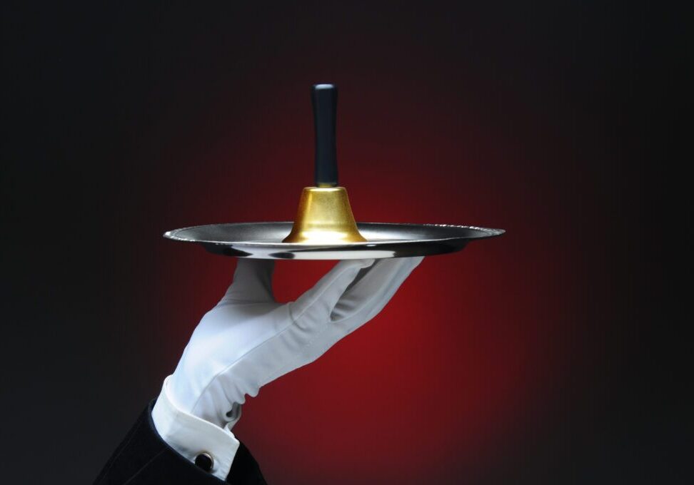 Closeup of a white gloved hand holding a service bell on a silver tray. Horizontal format on a light to dark red background.